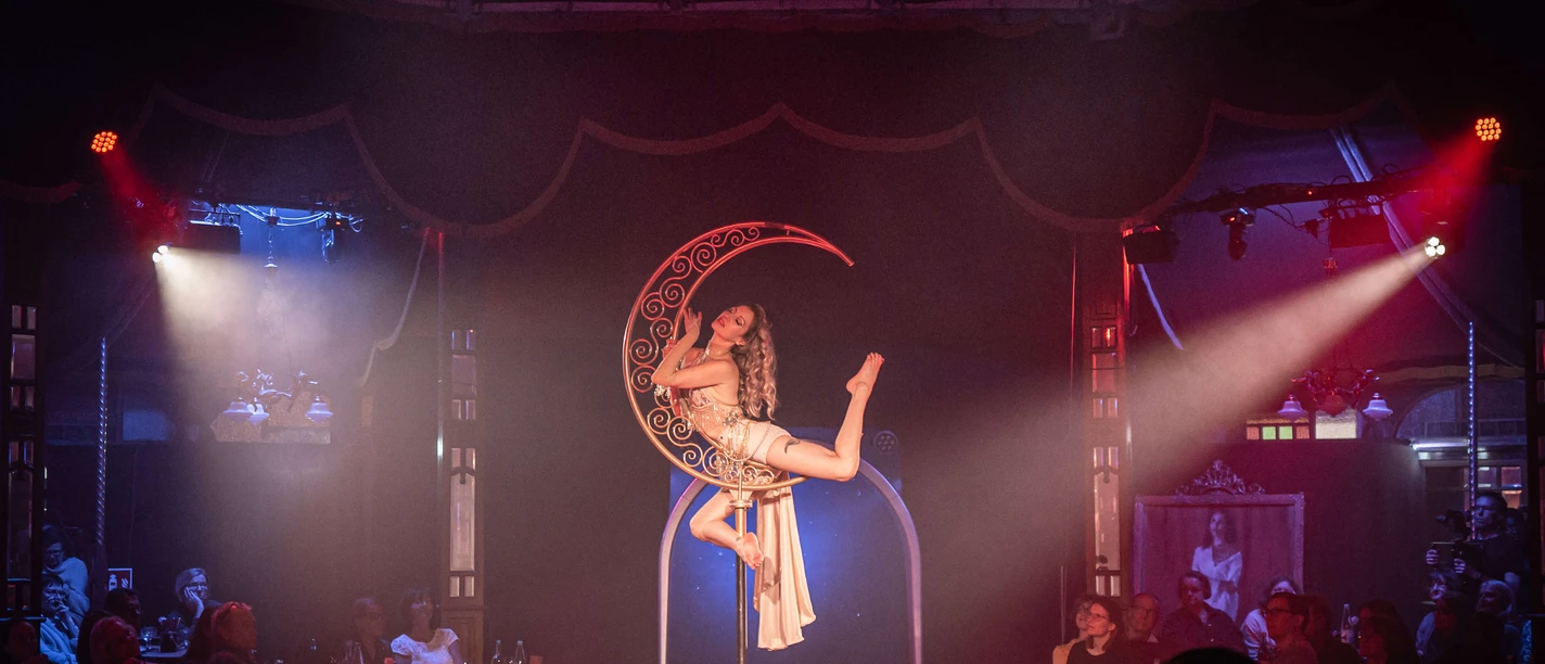 „Fly Me to the Moon – Sinatra meets Varieté“ Akrobatin posiert am beleuchteten Halbmond im Spiegelpalast vor Publikum.