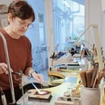 Eine Frau steht in einer Kunstwerkstatt vor einem Werktisch, auf dem viele verschiedene Werkzeuge und Materialien liegen, und fertigt ein Kunstobjekt an.