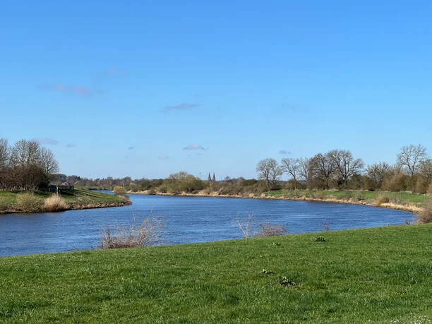 Die Weser mit Blick auf die Kirche in Bücken.jpg Grüne Ufer an der Weser unter blauem Himmel. In der Ferne ragen die Türme der Bücker Kirche auf.