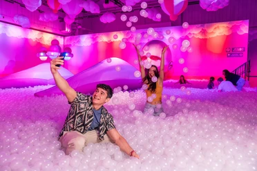 BUBBLE PLANET Menschen spielen in einem pink beleuchteten Bällebecken mit schwebenden Ballons