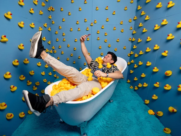 BUBBLE PLANET Verspielter Raum voller gelber Quietscheenten mit entspannter Person in einer Wanne beim Selfie