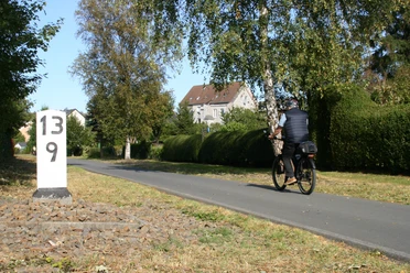 Auf den Spuren der Lenneper Tuchmacher Radfahrende Person auf asphaltiertem Weg neben einem weißen Kilometerstein, umgeben von Bäumen und einem Haus.