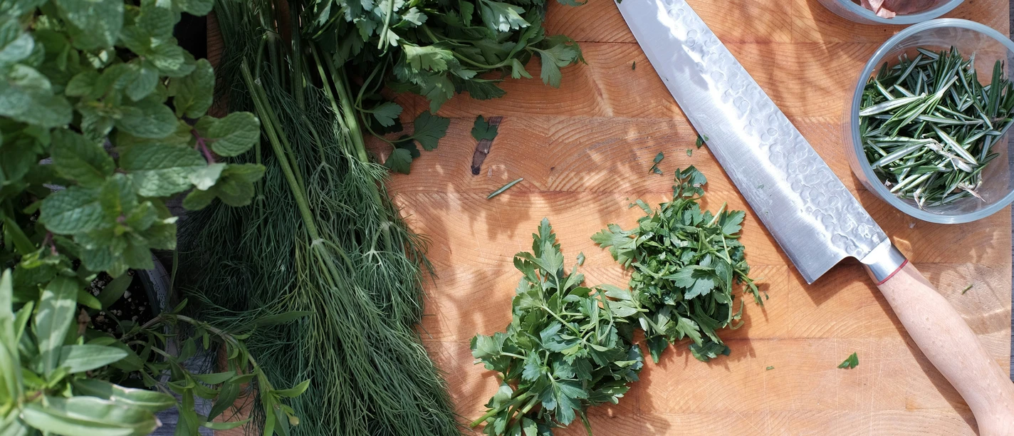 verschiedene Kräuter auf Holzbrett. Daneben liegt ein Messer, Zwiebeln und Knoblauch