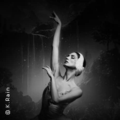 Ballerina in schwarzer Umgebung mit erhobenem Arm und federartigem Kopfschmuck in Pose.