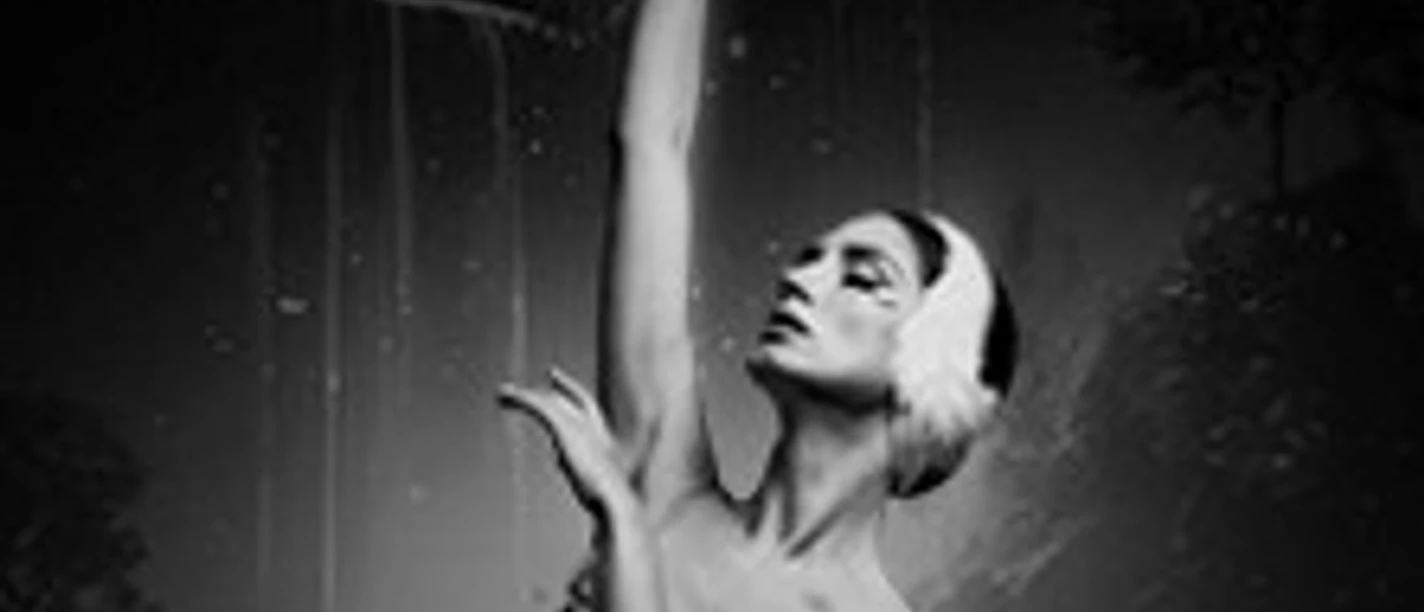 Ballerina in schwarzer Umgebung mit erhobenem Arm und federartigem Kopfschmuck in Pose.