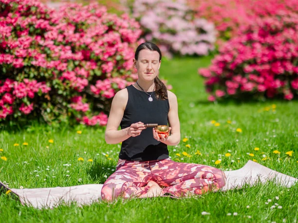 Meditation im Schneidersitz Frau sitzt im Schneidersitz auf einer Matte, meditiert mit einer Schale in der Hand, umgeben von blühenden Blumen.