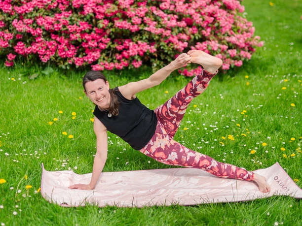 Frau in Yogahaltung auf einer Matte, umgeben von bunten Blumen und grünem Gras.