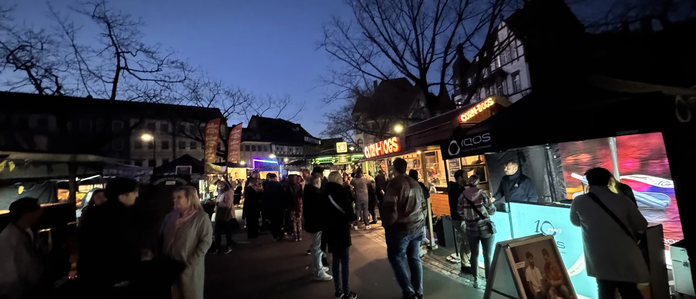 Happy Food Festival Menschen an beleuchteten Ständen auf einem abendlichen Street Food Festival vor Fachwerkhäusern.