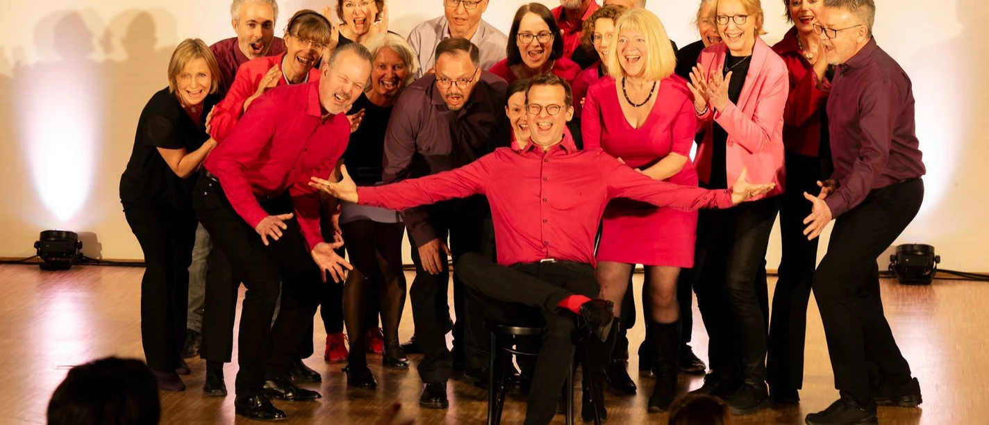 Auf einer hellen Bühne steht eine Gruppe von Männern und Frauen in schwarzer und roter Kleidung und posiert lachend für ein Gruppenfoto