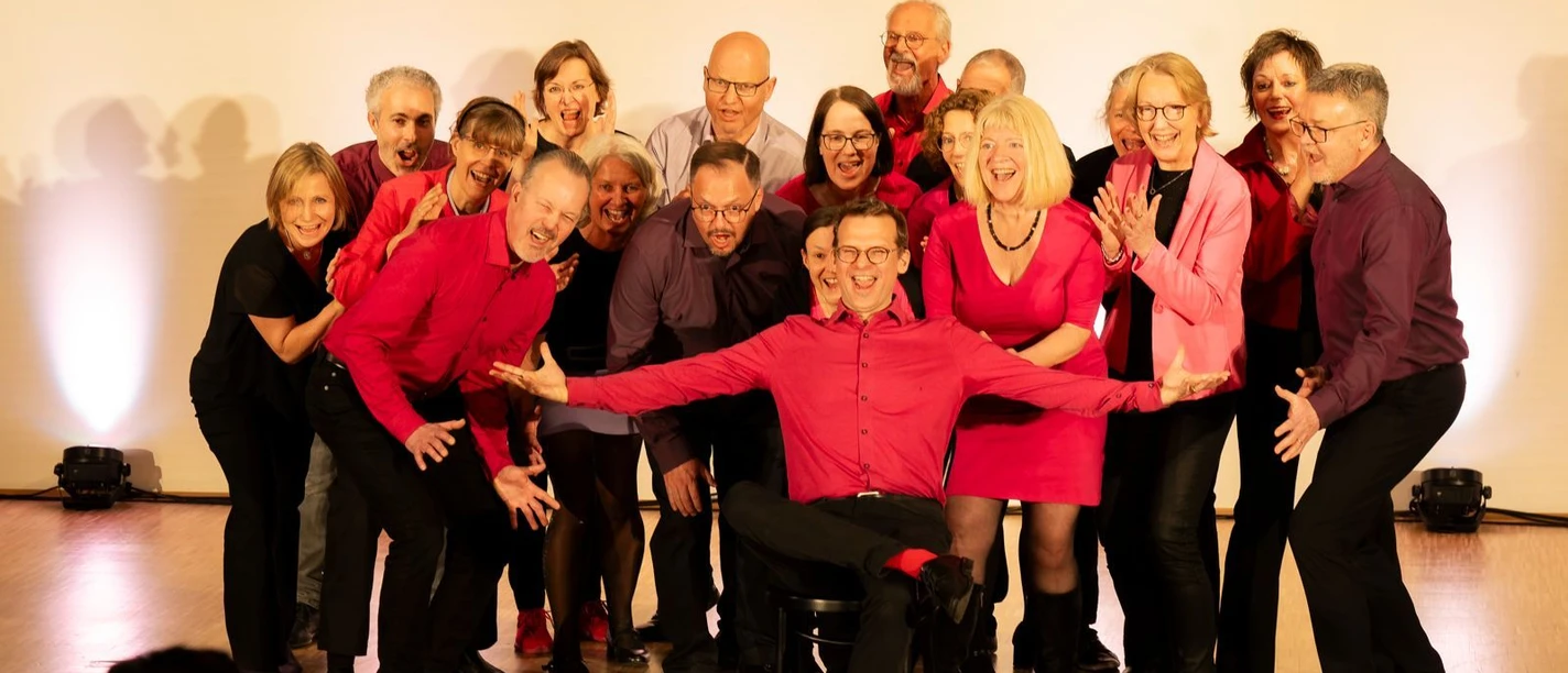 Auf einer hellen Bühne steht eine Gruppe von Männern und Frauen in schwarzer und roter Kleidung und posiert lachend für ein Gruppenfoto