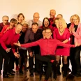 Chorkonzert frameless (c) frameless e.V..jpg Auf einer hellen Bühne steht eine Gruppe von Männern und Frauen in schwarzer und roter Kleidung und posiert lachend für ein Gruppenfoto