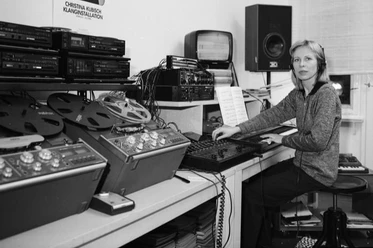 Ein schwarz-weiß-Bild zeigt eine Frau, die Kopfhörer trägt und vor Tischen mit Computern und Tonbändern sitzt.