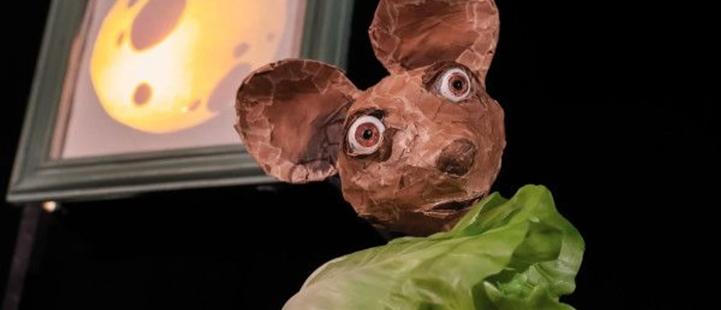 Die Puppenfigur einer Maus hält ein Salatblatt in der Hand. Im Hintergrund hängt das Bild eines Käsewürfels.