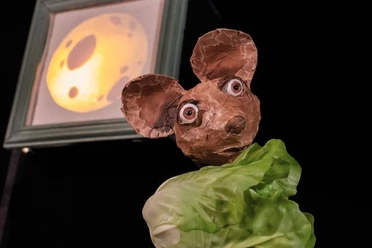Frederick_DAS DA Theater_Theater Starter (c) Sandra Hermannsen.jpg Die Puppenfigur einer Maus hält ein Salatblatt in der Hand. Im Hintergrund hängt das Bild eines Käsewürfels.