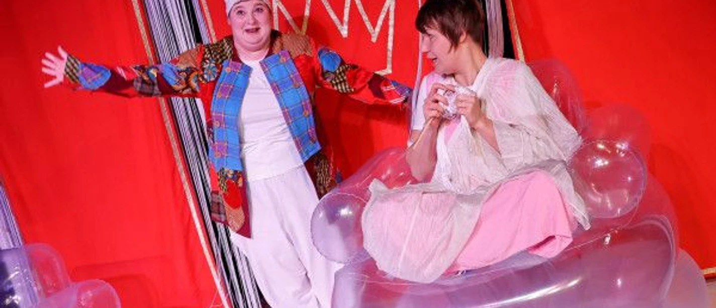 Zwei Schauspieler in farbenfrohen Kostümen stehen und sitzen auf einer Bühne mit rotem Hintergrund und schauen glücklich.