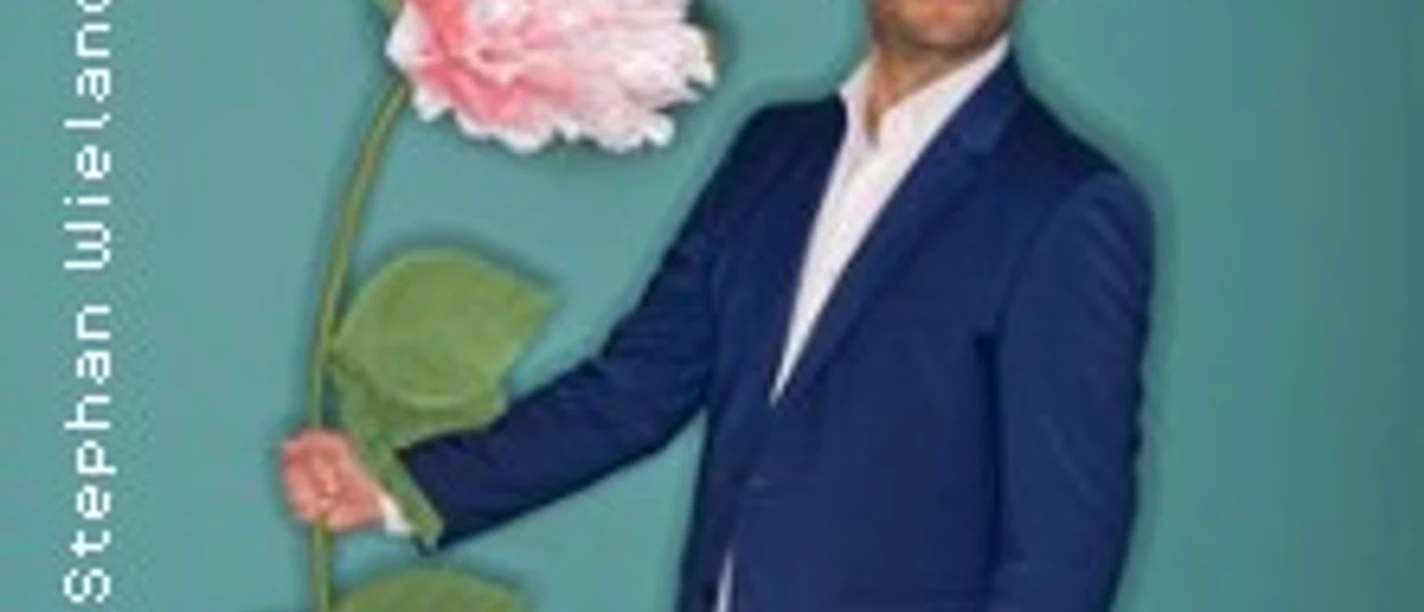 Mann im blauen Anzug hält übergroße rosa Blume vor türkisfarbenem Hintergrund