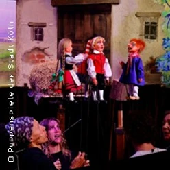 Handpuppen auf einer Bühne vor gemaltem Dorfhaus, darunter sichtbar spielende Darsteller.
