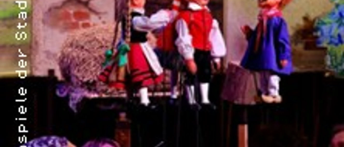 Handpuppen auf einer Bühne vor gemaltem Dorfhaus, darunter sichtbar spielende Darsteller.