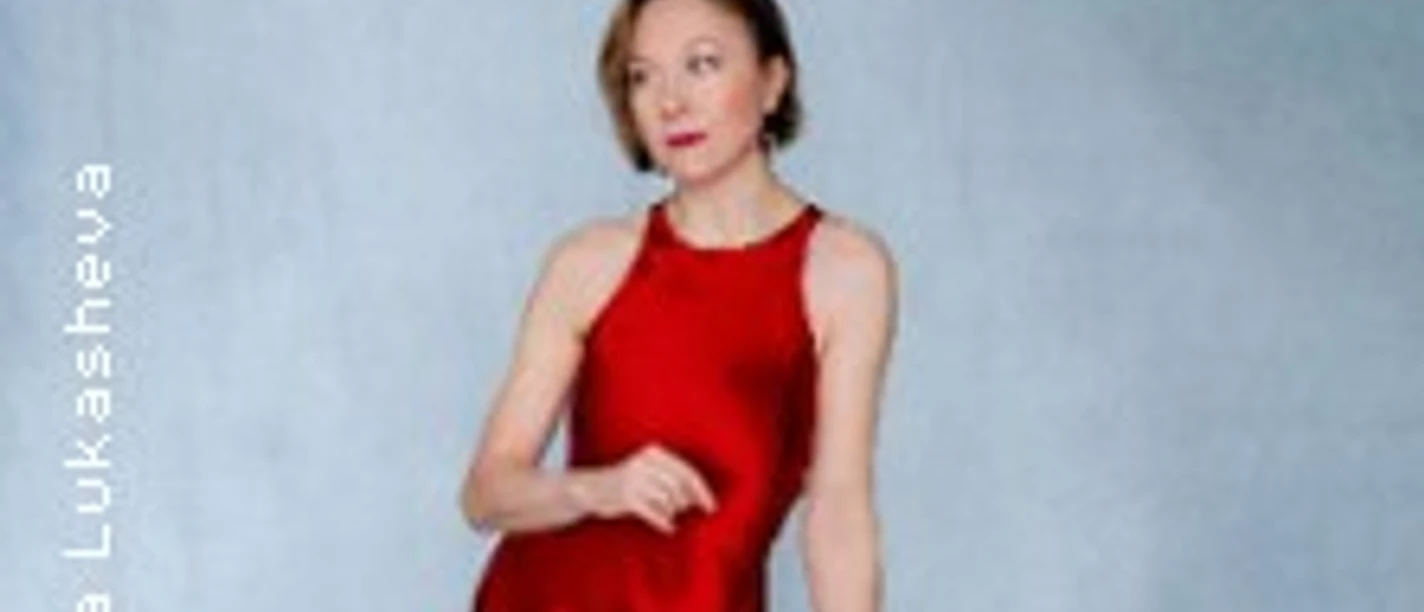 Eine Person in rotem Kleid lehnt an einem Hocker vor hellblauem Hintergrund.