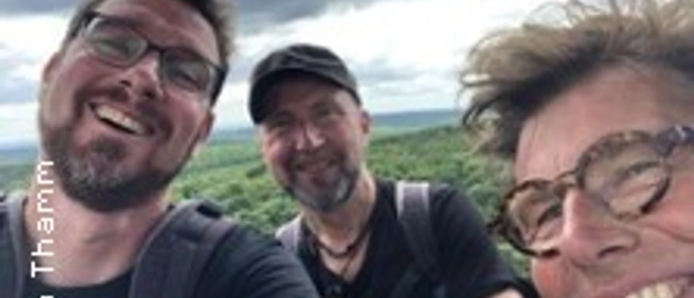 Drei lachende Personen vor bewölktem Himmel in grüner Landschaft