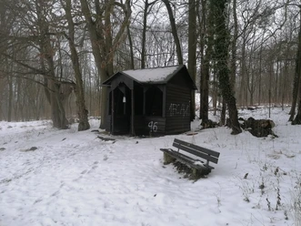 Königsstuhlhütte im Schnee Eine dunkle Holzhütte im verschneiten Winterwald. Davor steht eine leere Holzbank im Schnee.