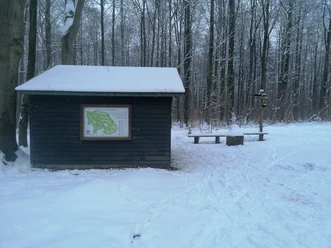 Schwarze Hütte im Schnee Dunkle Holzhütte mit Wanderkarte im verschneiten Wald, daneben Bänke und ein Wegweiser.