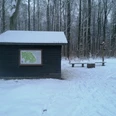 Schwarze Hütte im Schnee Dunkle Holzhütte mit Wanderkarte im verschneiten Wald, daneben Bänke und ein Wegweiser.