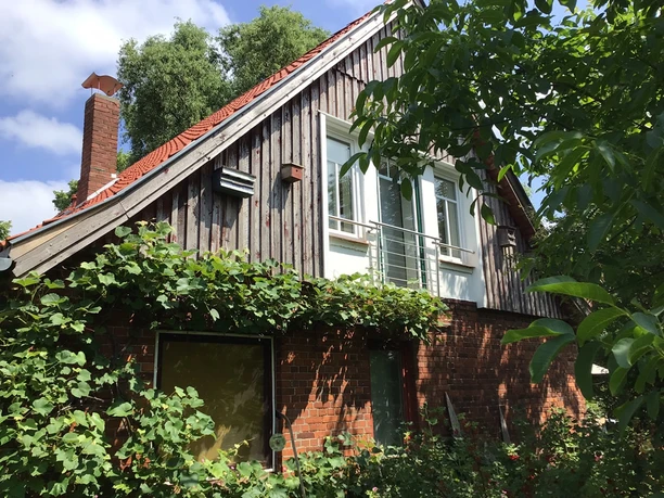 Suedgiebel Südgiebel eines Backsteinhauses mit Holzverkleidung und Balkon, umgeben von grünen Pflanzen.