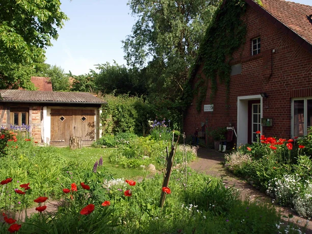 Vorgarten Eingang Ein Klinkerhaus mit grün bewachsenem Giebel und Holztür, umgeben von einem üppigen Blumengarten.
