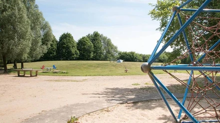 IMG_7254_WEB.jpg Klettergerüst und Tischtennisplatte auf Sandspielplatz mit angrenzender Rasenfläche und Bäumen ringsherum auf dem Spielplatz am Ginsterweg
