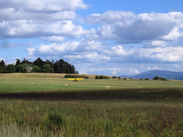 Flugplatz_Schaeferstuhl_Salzgitter_8_HD_16zu9.JPG Gelbes Sportflugzeug auf einer grünen Graspiste vor Bäumen und Bergen unter bewölktem Himmel.