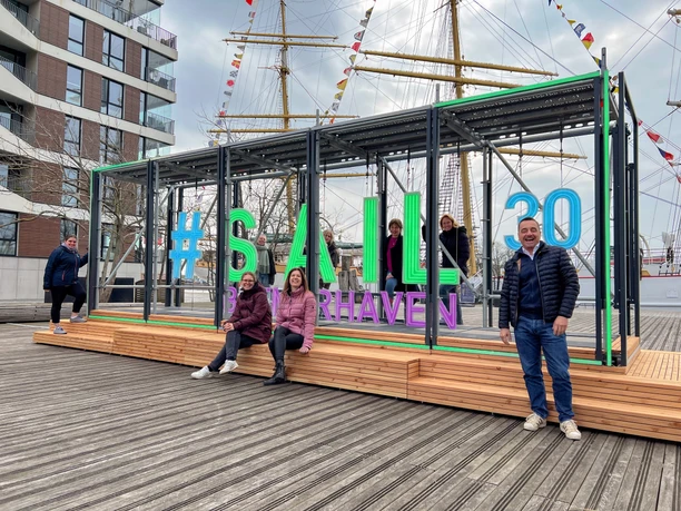 IMG_0362 (c) Thorsten Brockmann_Erlebnis Bremerhaven_leuchtend_2.png Fröhliche Gruppe am leuchtenden Schriftzug #SAIL30 Bremerhaven vor einem großen Segelschiff.