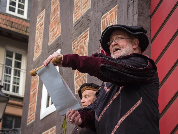 Verlesung der neuen Stadtrechte am Rathaus Sie sehen zwei Männer in historischer Kleidung vor dem Tor des historischen Frankenberger Rathauses stehen. Der Mann im Vordergrund trägt eine wichtige Verkündung von einer ausgerollten Papierrolle vor.t