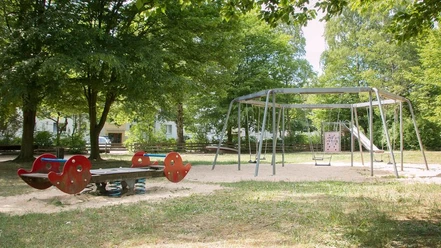 IMG_7244_WEB.jpg Wippe, Schaukel, Drehscheibe, Kletterwand und Rutsche umgeben von Bäumen auf dem Spielplatz Jakob-Kaiser-Straße