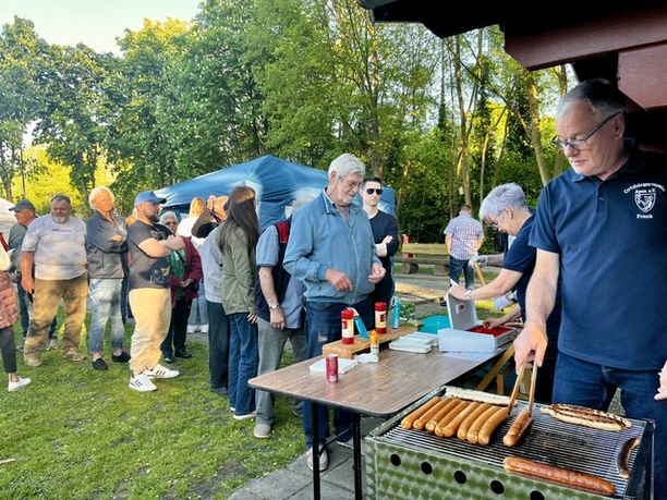Tanz in den Mai.jpeg Ein Mann grillt draußen Würstchen, während eine Menschenschlange davor wartet.
