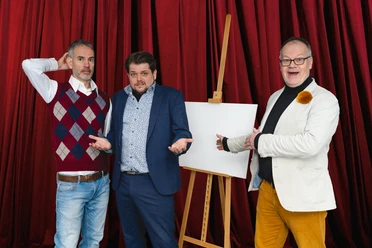 FotoBühnenstürmer.jpg Drei Männer gestikulieren vor rotem Vorhang neben einer leeren Leinwand auf Staffelei.
