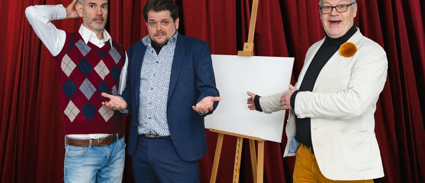 FotoBühnenstürmer.jpg Drei Männer gestikulieren vor rotem Vorhang neben einer leeren Leinwand auf Staffelei.