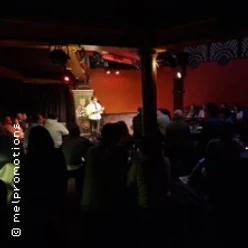 Bühne mit zwei auftretenden Personen, umgeben von sitzendem Publikum in gedimmtem Raum.