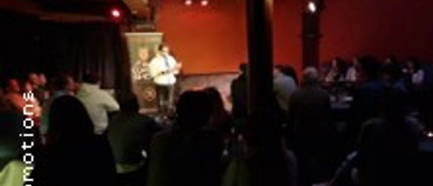 Bühne mit zwei auftretenden Personen, umgeben von sitzendem Publikum in gedimmtem Raum.