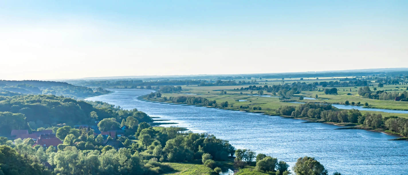 Elbekirchentag - An der Elbe.jpeg Die Elbe schlängelt sich sanft durch eine weite, grüne Landschaft unter blauem Himmel.