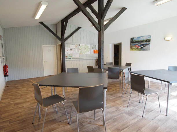 Innen Heller Aufenthaltsraum mit Holzboden, dunklen Holzbalken sowie grauen Tischen und Stühlen.