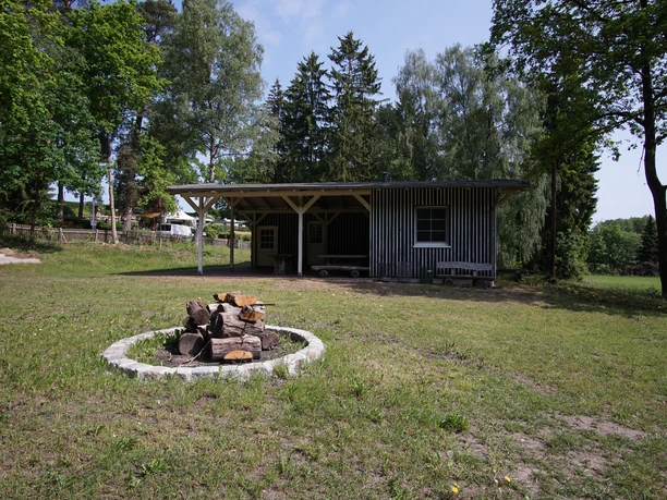 Feuerstelle.JPG Feuerstelle mit gestapeltem Holz auf einer Wiese. Im Hintergrund ein Holzgebäude unter Bäumen.