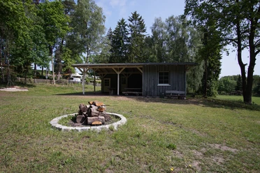 Feuerstelle Feuerstelle mit gestapeltem Holz auf einer Wiese. Im Hintergrund ein Holzgebäude unter Bäumen.