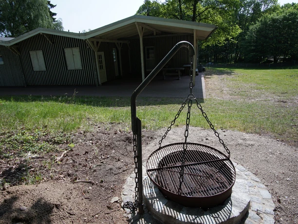 Grillstation Runder Schwenkgrill über einer Feuerstelle auf einer Wiese. Im Hintergrund ein Holzgebäude.