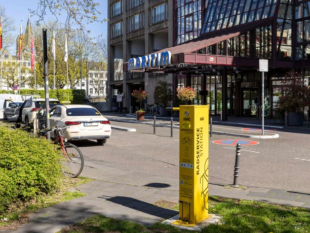 ADAC Radservice Station Gelbe ADAC-Radservicestation nahe Hoteleingang, umgeben von Autos, Fahrrädern und Grünflächen.