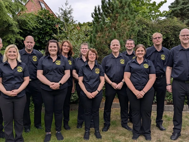 Falcon_Regiment.jpeg Elf lächelnde Personen in schwarzen Hemden mit Logo stehen zusammen in einem grünen Garten.