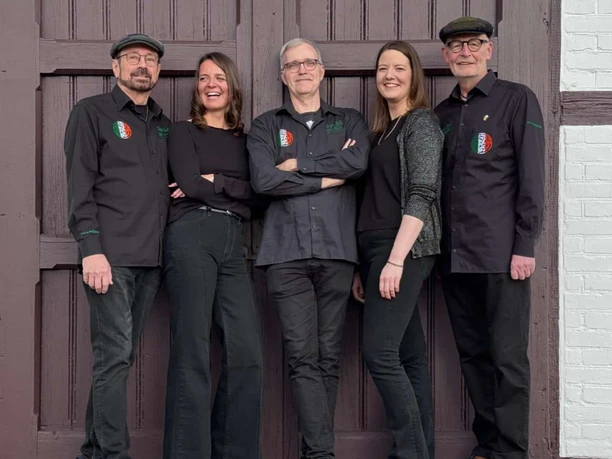 The_New_Foggy_Few.JPG Fünf lachende Musiker in schwarzer Kleidung posieren gemeinsam vor einem großen Holztor.