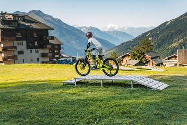 Bellwald Sommer skills park Ein Kind fährt mit einem Mountainbike über eine kleine Holzrampe auf einer Wiese vor alpiner Bergkulisse und umliegenden Gebäuden.