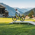 Bellwald Sommer skills park Ein Kind fährt mit einem Mountainbike über eine kleine Holzrampe auf einer Wiese vor alpiner Bergkulisse und umliegenden Gebäuden.