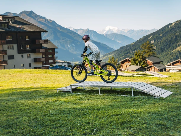 Bellwald Sommer skills park Ein Kind fährt mit einem Mountainbike über eine kleine Holzrampe auf einer Wiese vor alpiner Bergkulisse und umliegenden Gebäuden.